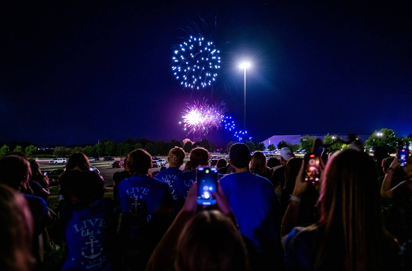 A crowd watches fireworks explode in the night sky, with some individuals capturing the moment on their phones.
